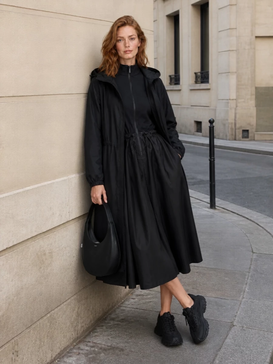 This look builds a monochrome, sport-utility column under the featherlight black Moncler parka: the technical zip-front midi dress and all-black trail sneaker read streamlined, functional and urban. The curved Coperni shoulder bag sharpens the silhouette with a graphic, architectural note, elevating the ensemble from practical rain shell to considered city uniform. Choosing the dress rather than separates keeps the lines clean and uninterrupted under the long outerwear, which is key to maintaining the minimalist, technical mood.
