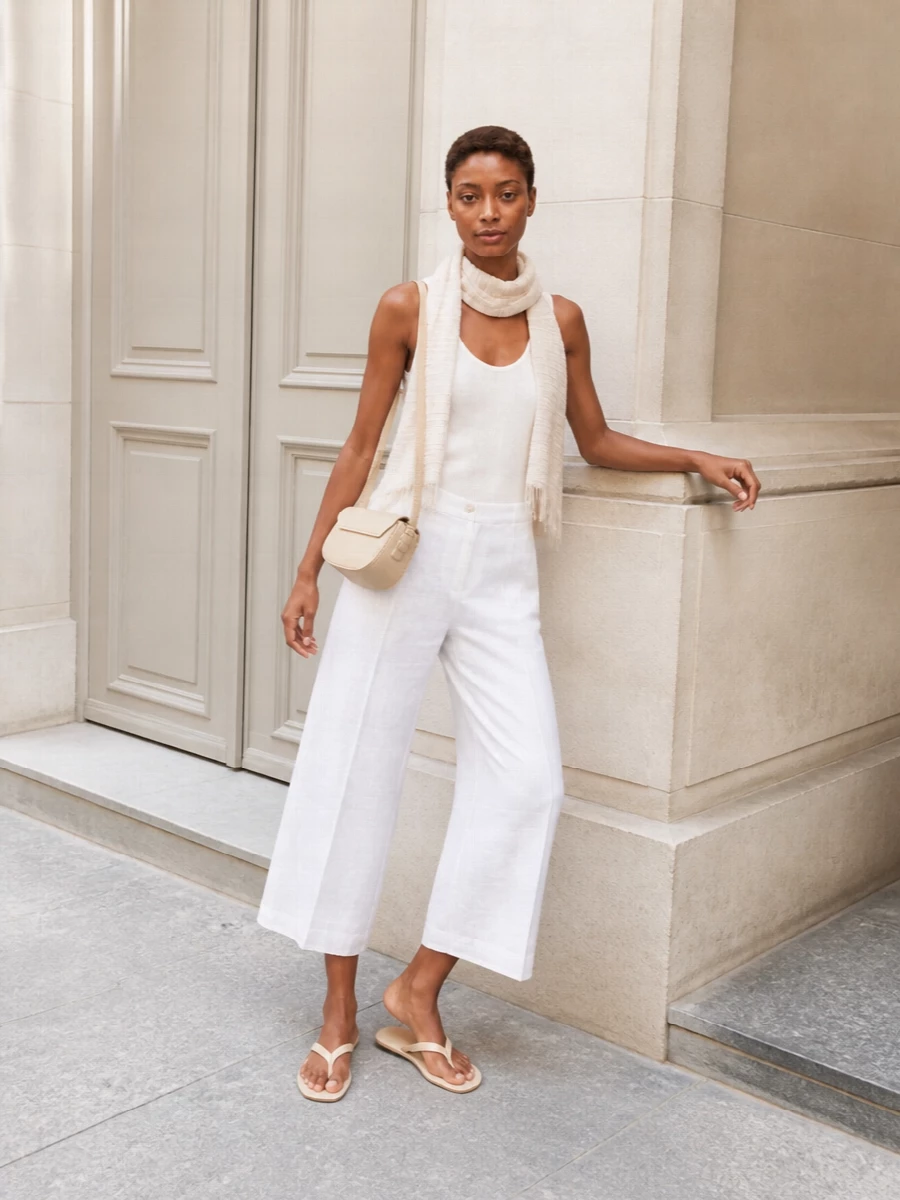A soft white ribbed tank meets cropped wide-leg trousers in the same airy palette for a calm, tonal take on warm weather dressing. Barely there cream thong slides and a buttercream half-moon bag keep everything light and precise, while a gauzy neutral wrap adds just a hint of texture without disturbing the quiet mood. The result is relaxed but polished enough for city days that drift into dinner.
