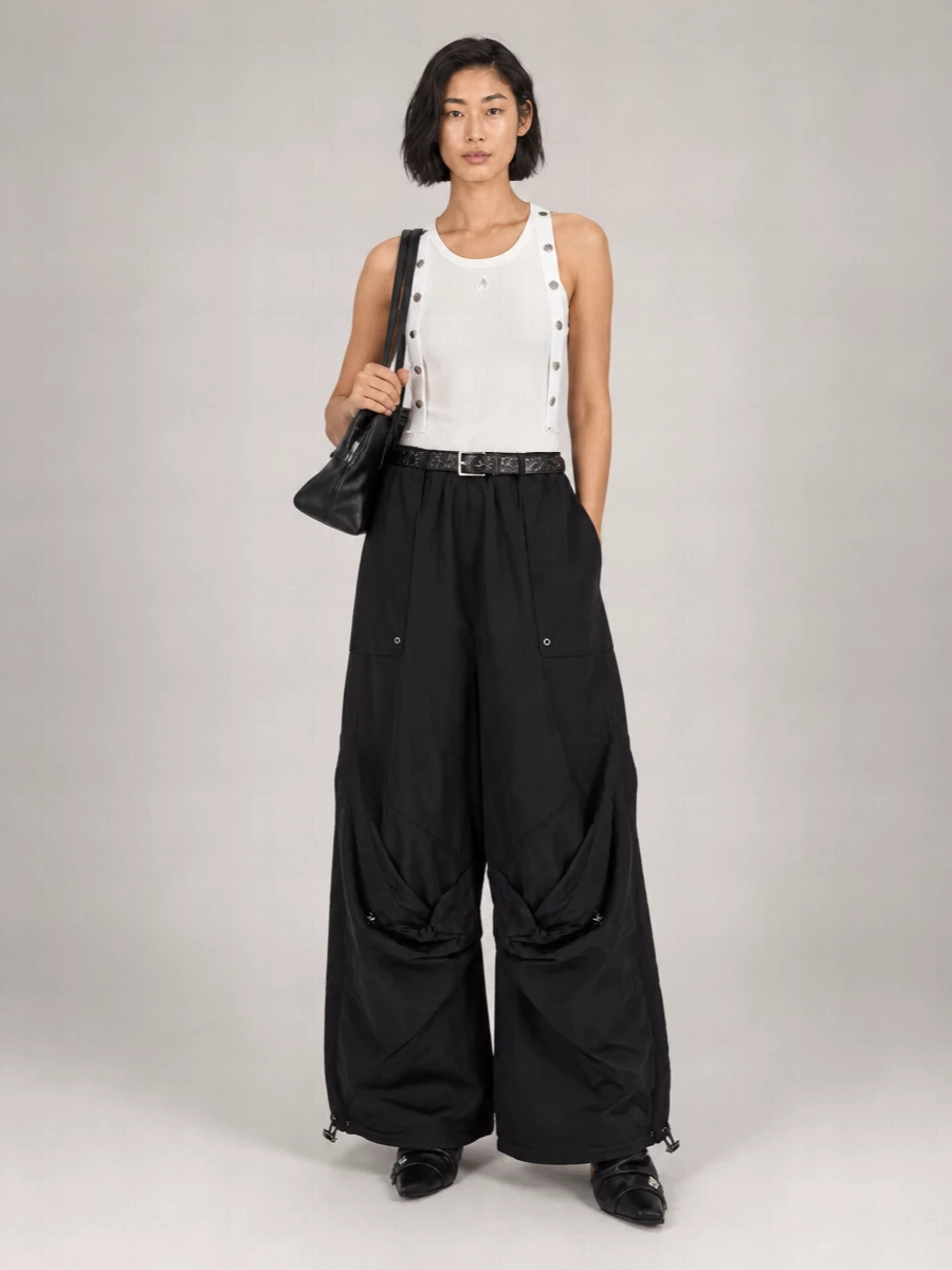 Ultra-wide nylon cargos sit high and dramatic against a lean white rib tank, keeping the palette tight and the attitude pure city street. Slouchy pointed black boots disappear under the sculpted hems, so the volume reads long and strong rather than heavy. A structured black shoulder bag and a slim belt with silver hardware finish it with just enough precision to feel intentional, not casual.