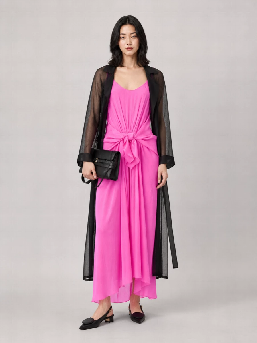 A shot of neon pink takes center stage, anchored by sharp black accessories that keep the drama focused on the dress’s sculptural drape. The pointed slingbacks and structured clutch streamline the line from hip to floor, reading modern and quietly expensive rather than overtly playful. It is a confident warm-weather cocktail ensemble that moves easily from gallery opening to late dinner.