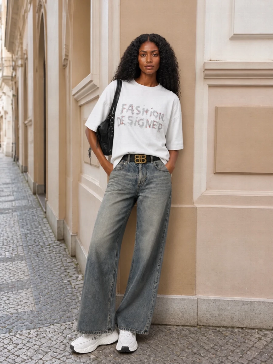 An oversized graphic tee falls over washed grey wide-leg denim for a quietly grungy, studio-off-duty attitude. Chunky monochrome runners keep the energy sporty and grounded, while a slouchy black moto tote and a slim BB belt add just enough hardware to feel intentional without breaking the nonchalant mood.