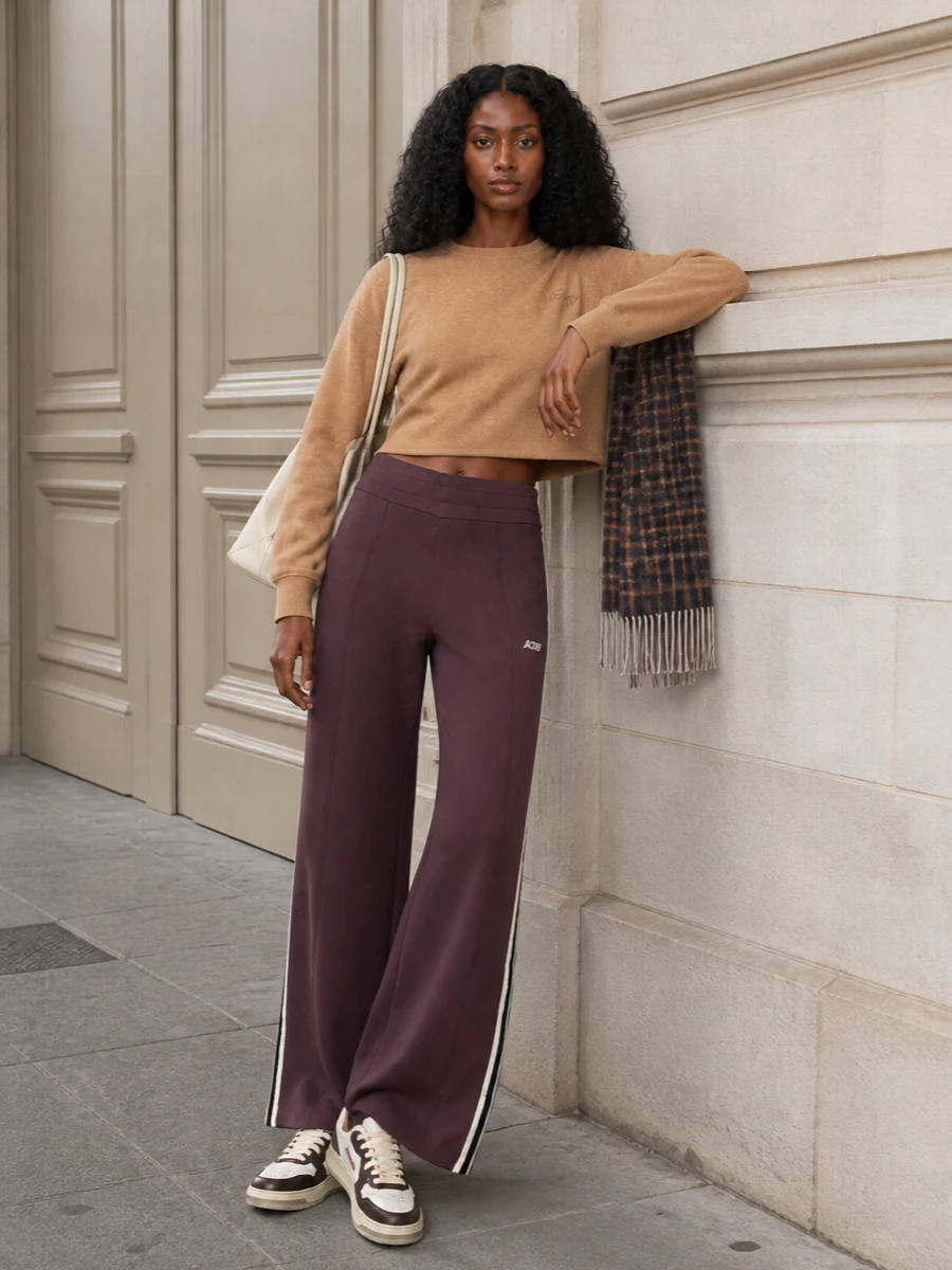 A tonal sport set built around the mauve-brown wide-leg track pant, grounded with a cropped camel sweatshirt that keeps the proportions sharp and relaxed. Vintage court sneakers echo the deep browns and soft whites, while the oversized cream tote and checked scarf add just enough texture for city days and travel. The result is retro-sporty, warm and quietly polished, ideal for off-duty errands or long-haul comfort that still feels intentional.