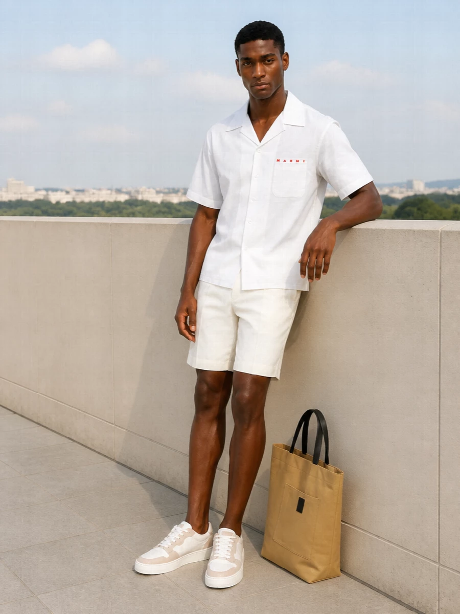 A crisp white camp collar shirt meets ivory tailored shorts for a monochrome summer uniform that feels precise yet relaxed. Soft off‑white court sneakers and a sand canvas tote keep everything in a low-contrast, quietly luxurious register. The result is warm-weather dressing that reads intentional from terrace coffee to late-afternoon gallery visits.