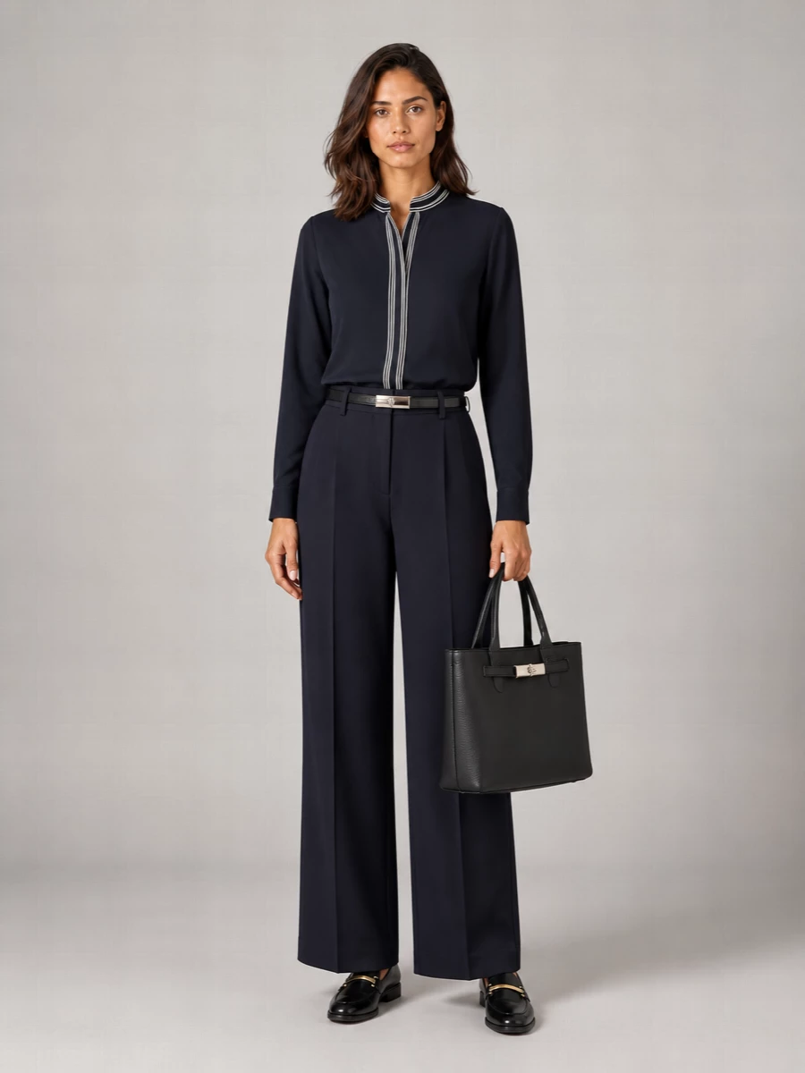 Deep navy tailoring stays sharp but not severe, with the contrast-trim blouse adding just enough graphic interest against the fluid high-rise trouser. Polished black loafers and a structured charcoal tote keep the mood disciplined and quietly expensive, while a slim belt finishes the long, leggy line without interrupting the minimalist ease.