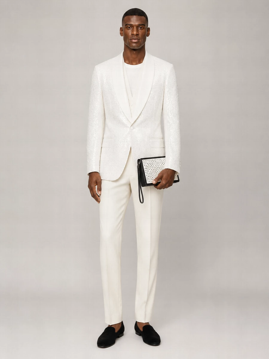 A white-on-ivory column of Dolce&Gabbana tailoring lets the sequin shawl jacket own the room, with tone-on-tone trousers and a clean single-breasted blazer keeping the silhouette razor sharp. Black velvet slippers and a graphic wristlet ground the light palette, adding just enough gravity for summer galas, terraces and destination weddings after dark. The result is formal, photogenic and unmistakably Italian without spilling into costume.