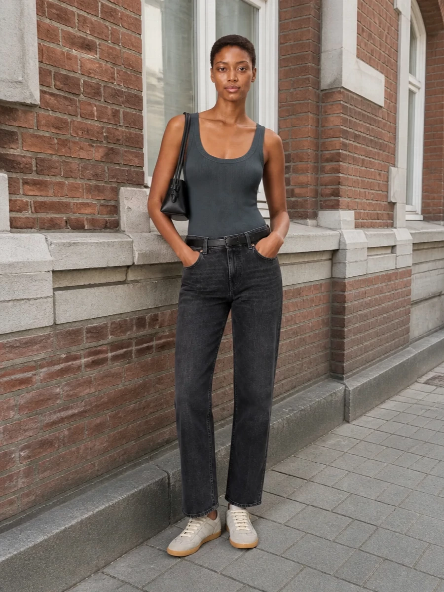 Washed charcoal denim is paired with a sculpted ribbed tank for a clean, tonal base that feels relaxed but precise. Grey suede trainers and a small black shoulder bag keep everything grounded in quiet luxury, while the slim belt adds just enough structure at the waist. It is an easy city uniform that reads considered from brunch to late walks home.