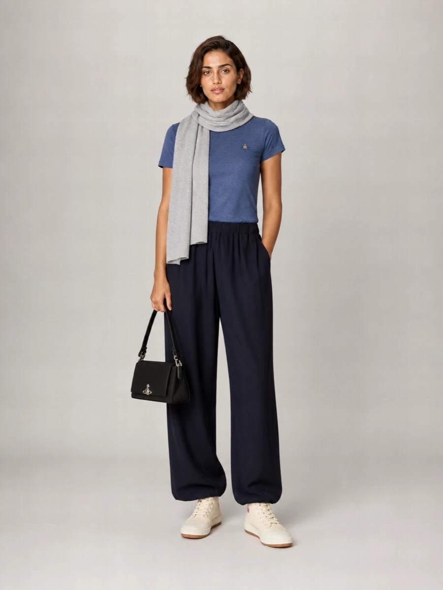 A slate Orb tee pairs with fluid pull-on navy trousers for a clean, relaxed line that feels considered but unfussy. Cream high-tops keep it casually grounded, while a compact black baguette and soft grey scarf add just enough structure and texture for city days. The result is quietly branded streetwear with a preppy, minimal twist that works from coffee runs to low-key dinners.