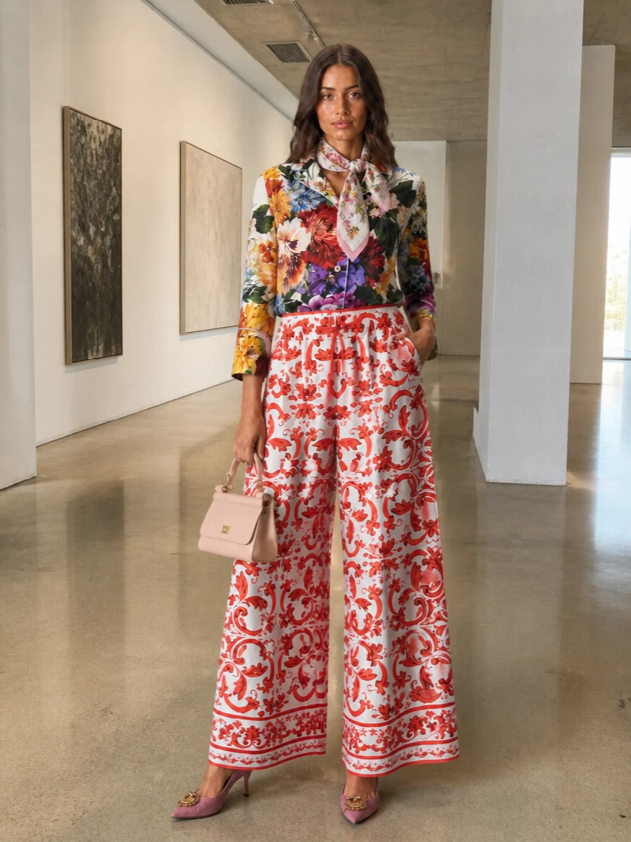 The floral pajama shirt meets the red baroque wide-leg trouser in a romantic, tailored take on coordinated prints, all grounded by supple silk textures and a precise waistline. Dusty rose heels and a matching blush top handle keep the mood soft and Italian, adding height and polish without stealing attention from the statement pairing. A delicate floral foulard finishes the story with a quiet nod to couture garden references.