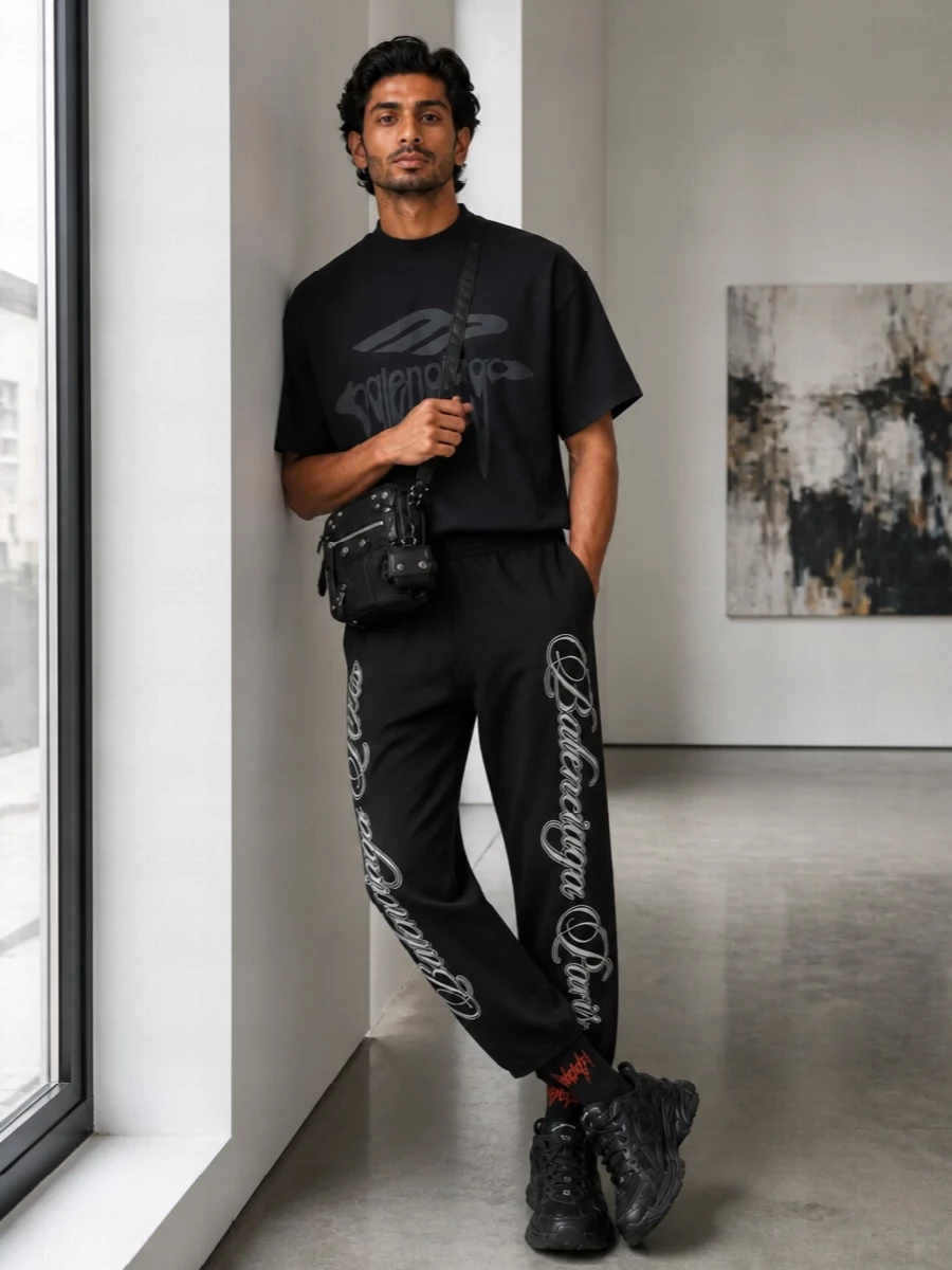 This is a pure Balenciaga street set built on a washed black, logo‑to‑logo conversation between tee and track pant. The all‑black technical runners and moto crossbody keep the mood sharp and streamlined, while the graphic socks add just enough irreverence peeking out above the sneakers. The result is relaxed in fit but very intentional in how the branding and textures stack from shoulder to sole.