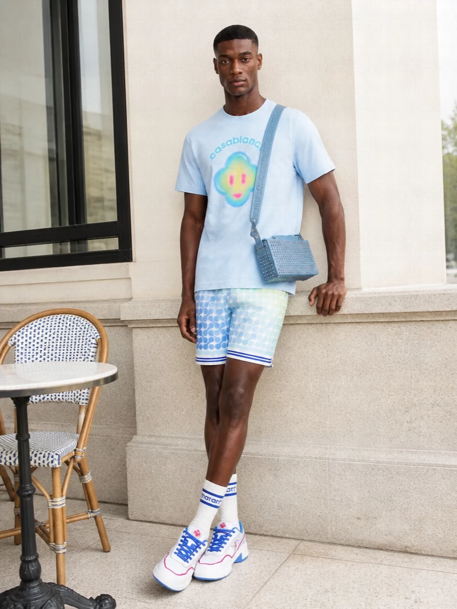 A soft-sunrise palette runs through this outfit, with the hazy sky-blue tee sitting over ombré deck shorts in matching icy blues and lime for a light, resort-ready base. Clean white and cobalt court sneakers keep the energy athletic and nostalgic, while the powder-blue studded crossbody and striped socks echo the colour story in a sharper, streetwear register. The result is a playful but considered warm-weather set that feels equally at home in the city or by the pool bar.