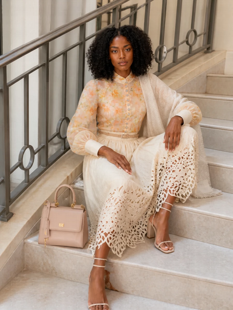Zimmermann’s sheer floral blouse meets a buttercream embroidered midi for a soft, high-waisted column of light that feels romantic yet composed. Champagne-gold sandals and a blush structured top-handle keep the mood polished for warm garden parties or terrace dinners, with a gauzy neutral stole as a barely-there layer around the shoulders. The result is luminous, feminine and quietly statement-making without a single hard edge in the palette.