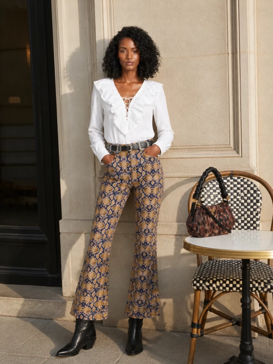 High-rise snake-print flares set the tone, balanced by a clean white ruffled blouse that keeps the print feeling intentional rather than chaotic. Black western boots and a slim double-buckle belt sharpen the 70s silhouette and ground the palette. A sculpted chocolate animal-print shoulder bag finishes it with compact, after-dark energy that still feels easy for city days.
