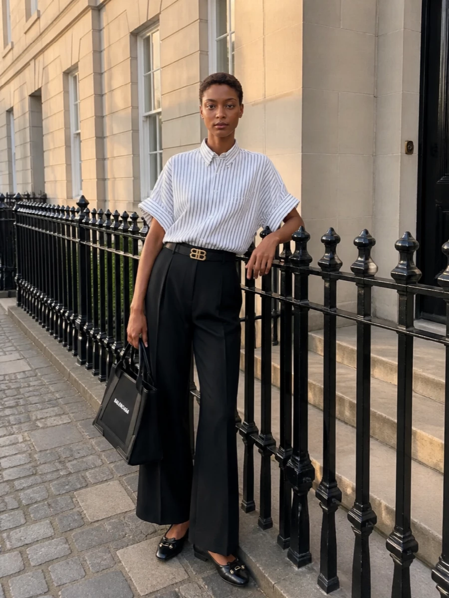 A striped, boxy shirt falls over fluid high-waisted black trousers for a long, quiet silhouette that feels smart without being formal. Sleek black flats and a structured monochrome bag keep the lines clean and graphic. A slim logo belt adds just enough definition to underline the Balenciaga attitude without breaking the ease.