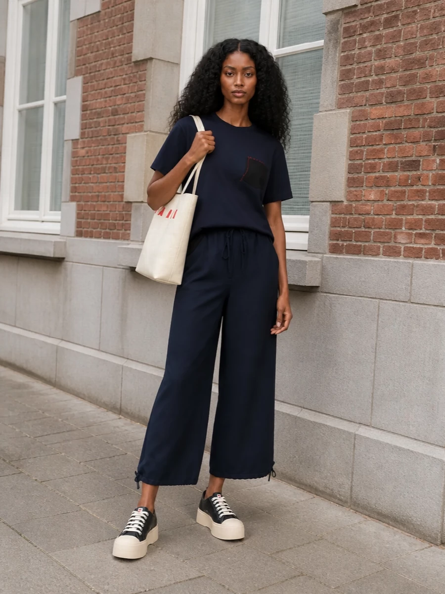 Navy on navy keeps this combination clean, letting the sculpted tee and cropped wide-leg trouser do the work in proportion and line. Platform sneakers and a structured cream tote introduce just enough graphic contrast, with hits of red linking tee and bag for a quietly coherent, street‑leaning finish. The result is relaxed but intentional, ideal for city days where comfort still reads considered.