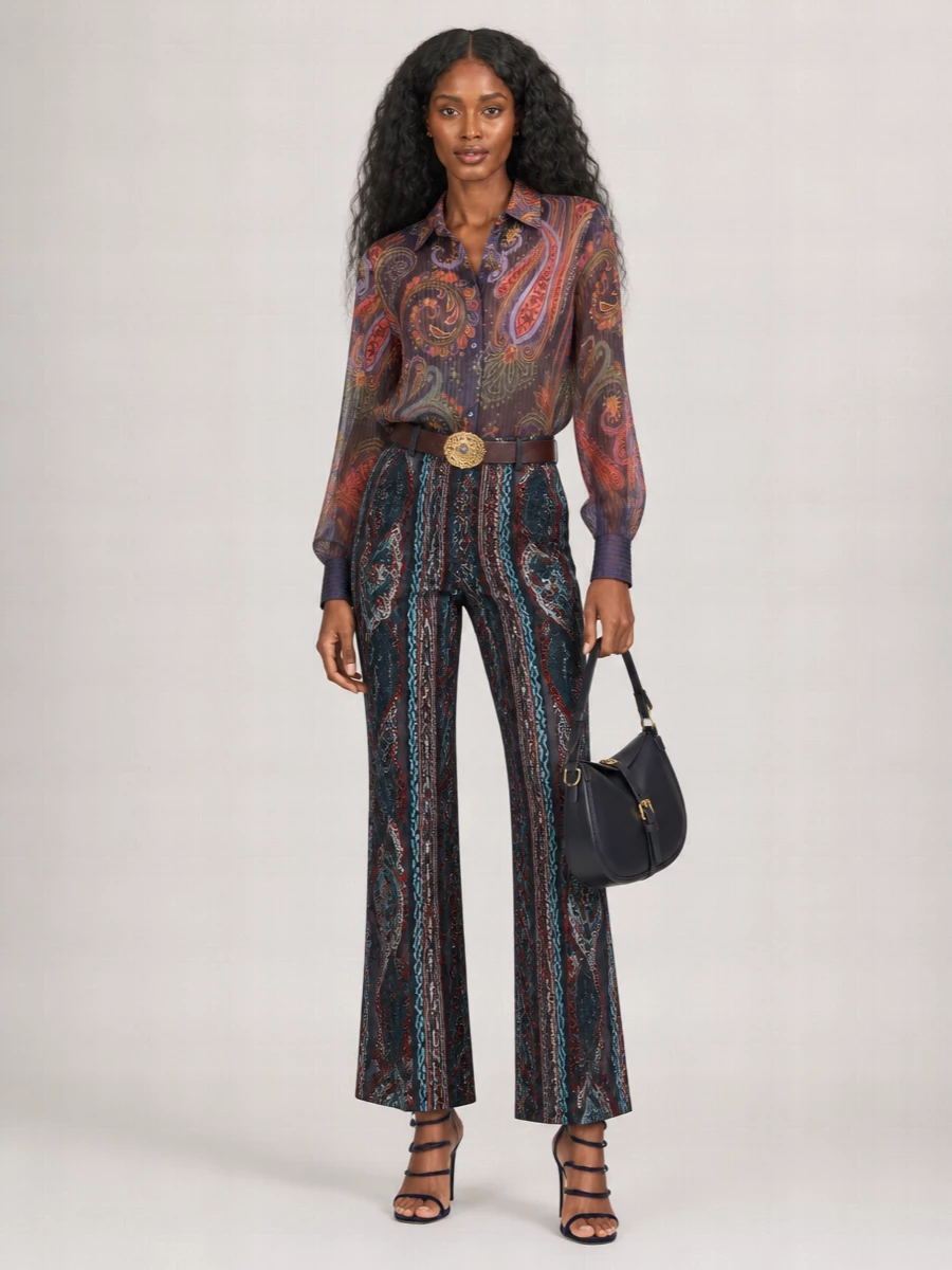 An inky paisley-on-paisley pairing turns the high-waisted flares into a rich evening statement, with the sheer shirt echoing their print while keeping the upper body soft and light. A sculptural navy spiral sandal and structured navy saddle bag lengthen and streamline the silhouette, so the pattern play feels intentional rather than busy. The jeweled belt is the final touch, cinching the waist and quietly picking up the trousers’ fiery tones without stealing focus.