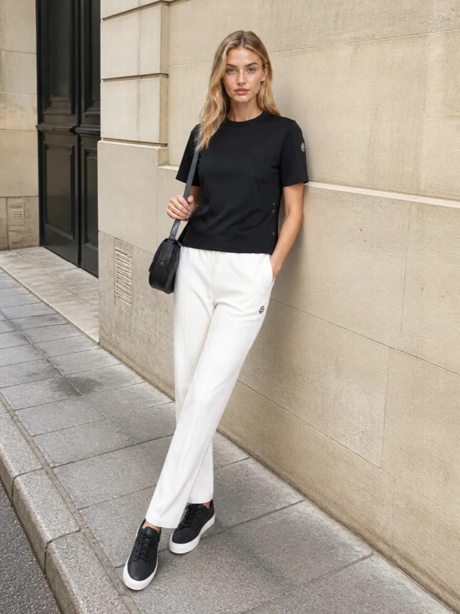 A boxy black Moncler tee is paired with off‑white straight-leg Moncler track pants for a graphic yet softly relaxed monochrome base. Black-and-white Moncler sneakers keep the line sporty and streamlined, while the Celine crescent bag adds a refined, minimal touch that reads effortlessly polished for off-duty city days or travel.