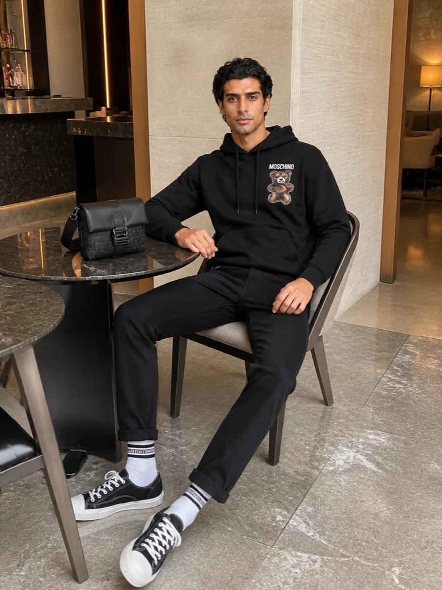 A graphic Moschino hoodie meets deep black straight-leg denim for a clean, street-led base that keeps the teddy detail in focus. Canvas low-tops and striped sport socks reinforce the monochrome, casual attitude, while the Fendi crossbody brings a compact, structured hit of luxury that still feels easy for daily wear. The result is a relaxed, city-ready outfit that balances playful branding with low-key polish.