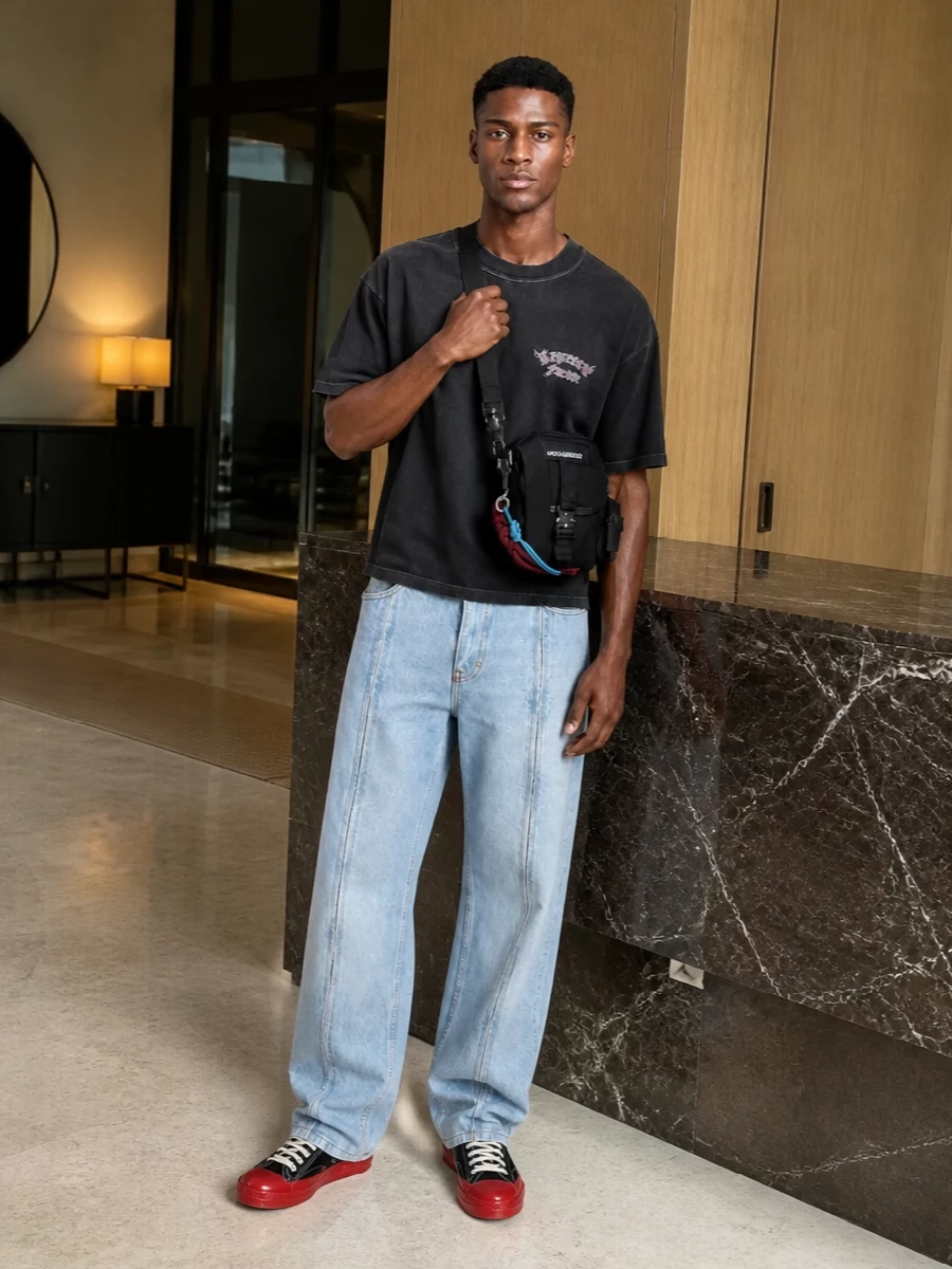 Washed black and light stonewash keep this street set relaxed, with the wide-leg denim pulling the tee into a slightly Y2K, skate-adjacent space. The black and red-accented sneakers ground everything in a clean court profile, while the compact crossbody adds just enough hardware and function without disturbing the drape.