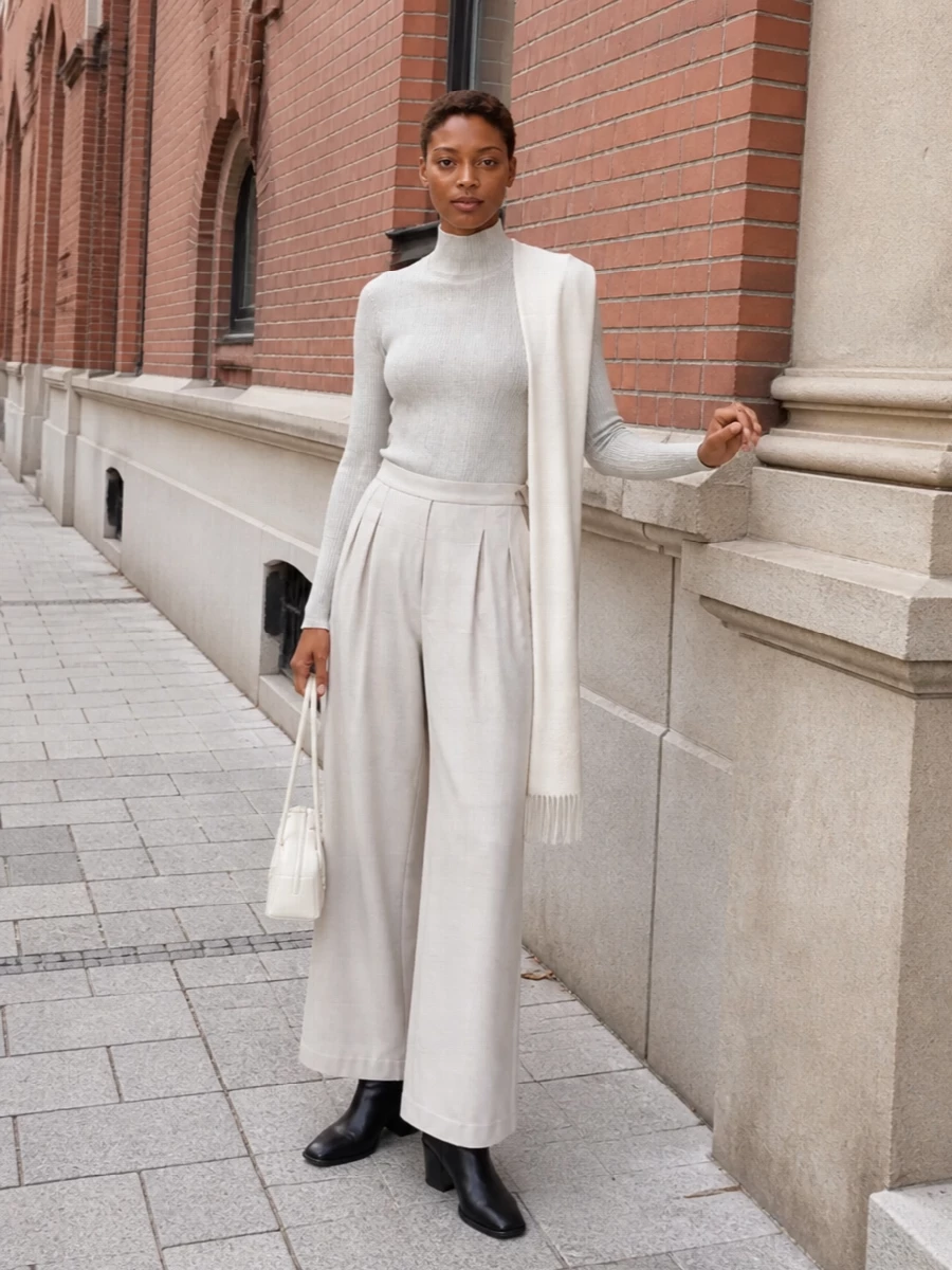 A column of pale stone and soft silver-grey creates a calm, elongated line that feels both precise and relaxed. The slim ribbed knit tucks neatly into the fluid high-waisted trouser, letting texture and proportion do the work. Black block-heel ankle boots and a sculpted ivory shoulder bag give the outfit quiet definition without disturbing its light, minimalist mood.