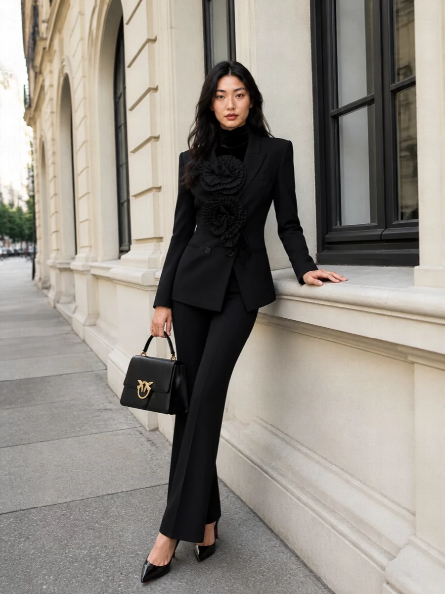 A sharply tailored black blazer and elongated trouser create a dark, romantic column that lets sculpted rosettes read as couture rather than costume. Glossy black stilettos and a structured top-handle bag keep the mood refined and city-ready, while a touch of velvet at the neck deepens the monochrome drama for evening dinners or late gallery openings.