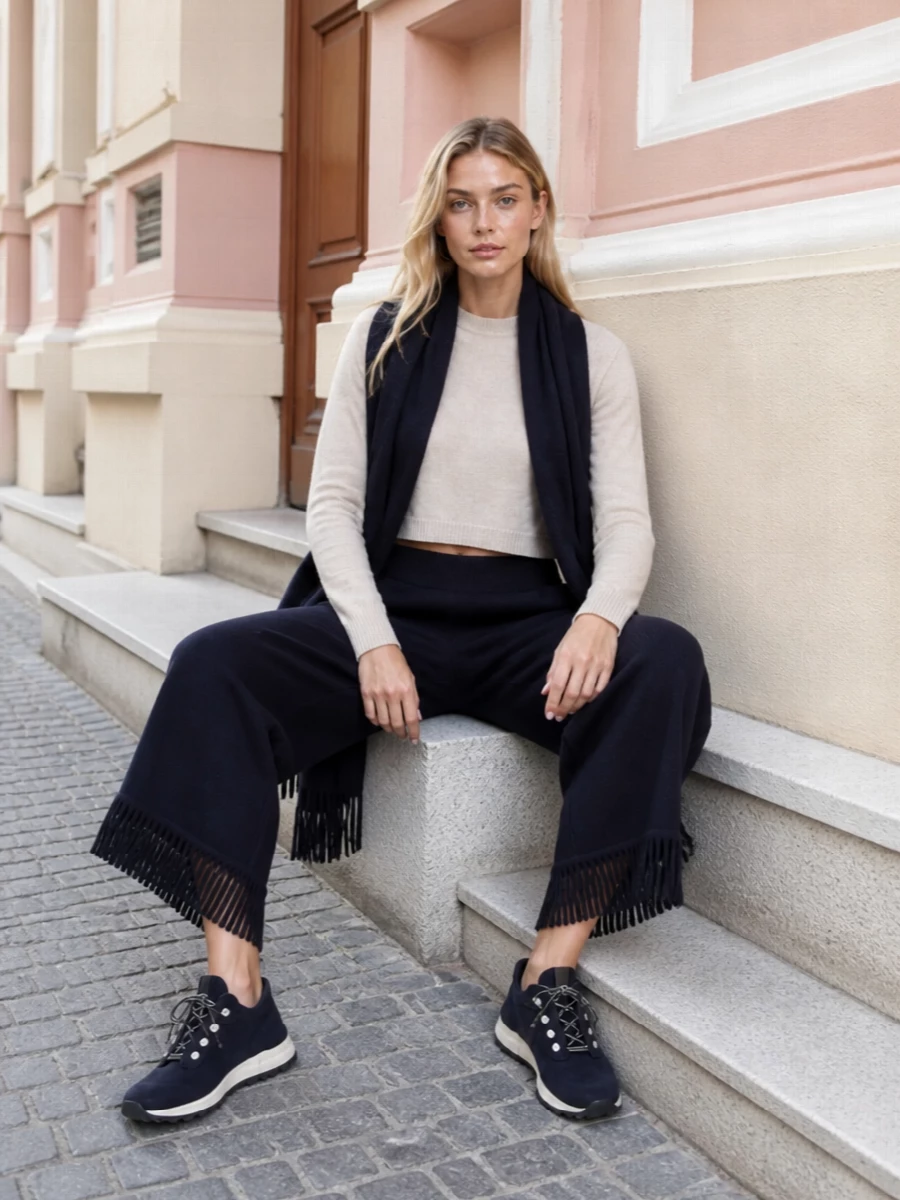 A cropped Alaïa knit in soft beige sits neatly on the high waist of Alanui’s navy fringe pants, keeping the proportions short over wide for a calm, modern line. Navy knit sneakers and a structured Alaïa clutch stay in the same deep palette, so the texture and silhouette do the talking rather than colour contrast. A fluid ink scarf finishes the outfit with a vertical, cocooning note that still feels considered and polished.