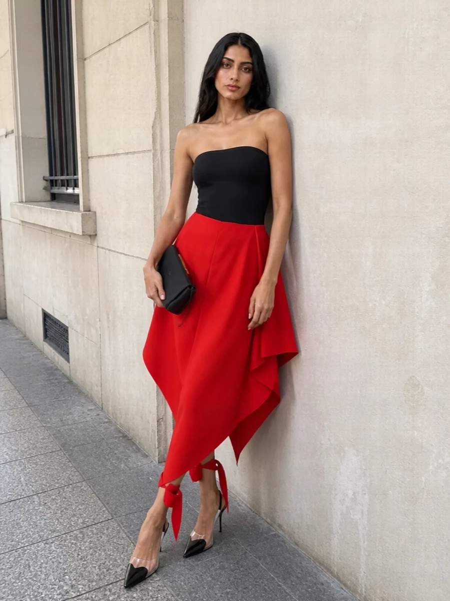 This look pairs the sculpted red Alaïa skirt with a pure, strapless black bodysuit for a graphic, body‑skimming top line against fluid, asymmetric volume. Black patent‑and‑clear slingbacks and a razor‑slim black clutch keep the accessories sharp yet understated, reinforcing the architectural mood while letting the vivid red drapery command full attention. The result is a modern cocktail silhouette that feels both disciplined and deeply feminine.