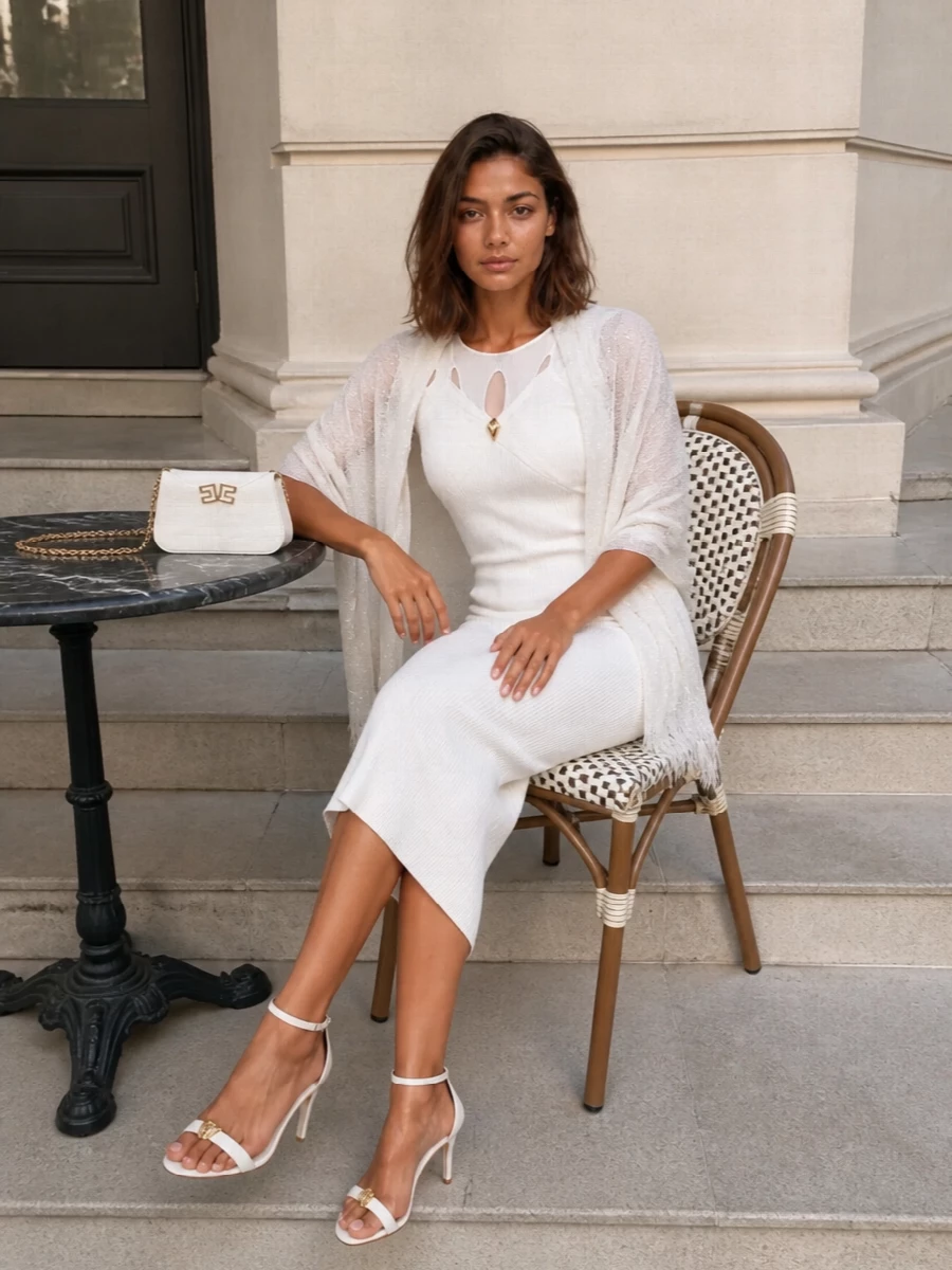 A clean white column in ribbed knit is framed with equally refined accessories, all playing in the same soft ivory and gold register. Fine straps, a compact baguette and a gossamer wrap keep the silhouette long and uninterrupted, letting the sheer yoke and charm at the neckline stay quietly in focus.