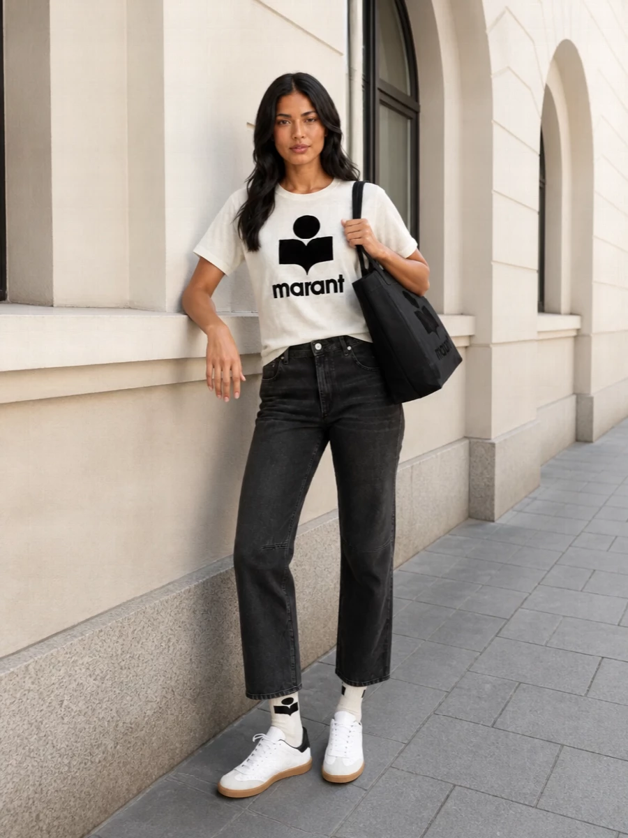 A soft white logo tee meets washed charcoal denim for a clean, graphic take on Parisian street style. White leather sneakers and an oversized black tote keep the palette tight and practical for city days. Logo socks peeking above the gum soles add just enough attitude while staying quietly polished.