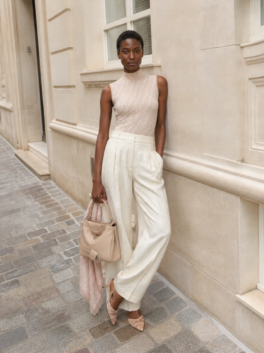 This look leans into Fendi’s quiet logo language with the sheer monogram mock-neck tucked into buttercream jacquard wide-leg trousers, creating an elongated, tailored silhouette in soft, sunlit neutrals. Nude-blush mesh kitten-heel slingbacks and a sculptural nude-beige top-handle bag keep the mood polished yet effortless, ideal for summer dinners or refined city cocktails. A blush jacquard wrap adds a subtle rose glow without disturbing the clean, tonal harmony.