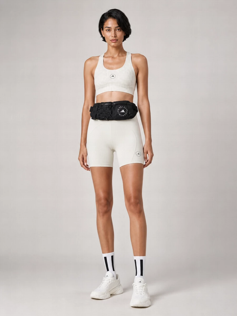 A monochrome training set built around ivory bike shorts and a soft white performance bra keeps the silhouette lean, sculpted and genuinely studio-ready. Clean white runners and graphic striped socks extend the light, minimal palette, while a black technical belt bag picks up the logo accents and adds hands-free practicality without disturbing the calm lines.