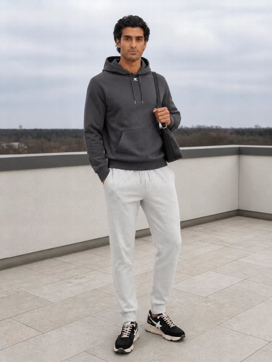 A charcoal hoodie and light grey joggers frame the sneakers in a soft monochrome gradient that feels precise but relaxed. The proportions stay lean and clean, so the chunky runner reads intentional rather than bulky. A compact black shoulder bag keeps everything in the quiet luxury lane without distracting from the line of the outfit.