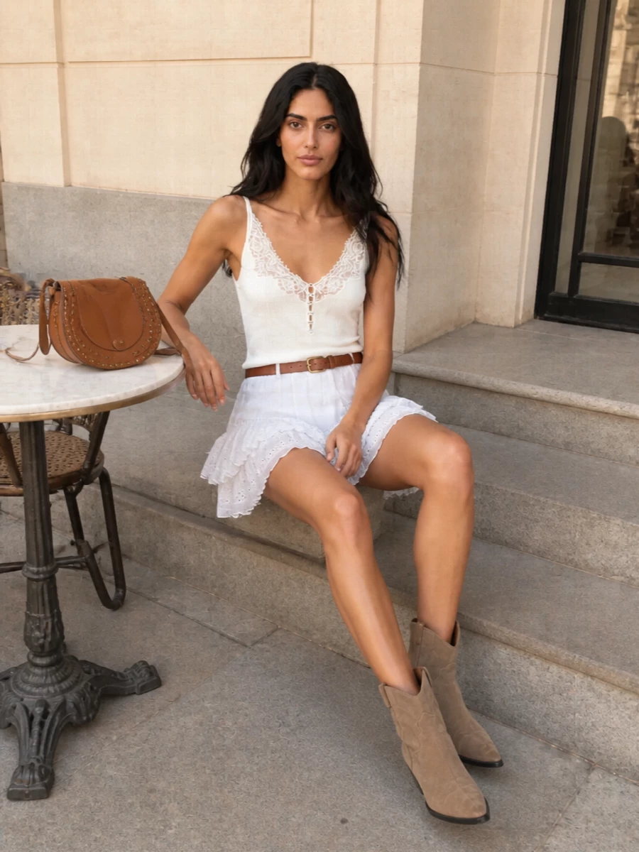 Softly romantic and quietly boho, this pairing lets the embroidered tiered mini do the talking, framed by a delicate lace camisole that skims the body. Taupe Western boots and a cognac saddle bag add grounded warmth and a Paris-meets-Riviera ease, while a slim leather belt gently defines the waist for a flattering, feminine line. It is an easy day-to-dinner outfit for late summer into early autumn, bare armed now and ready to layer later.