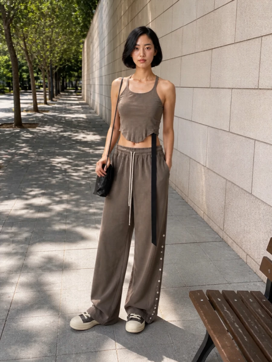 A washed taupe tank and matching wide-leg drawstring trouser create a long, fluid Rick Owens Drkshdw column in soft neutrals, sharpened by the asymmetric tie and snap‑side detailing. Chunky taupe low-top sneakers keep the palette continuous and street-led, while a slim black pouch adds just enough hardware to underline the utility mood without breaking the flow.