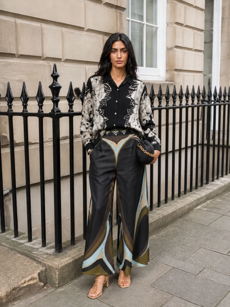 A dark, graphic Pucci maxi anchors the outfit, paired with a monochrome floral silk-feel shirt for a romantic yet composed take on print-on-print. Champagne-gold sandals and a softly ruched black clutch bring shimmer and polish without disturbing the moody palette. The result is boho-leaning cocktail dressing that feels fluid, modern and ready for terraces or gallery evenings.