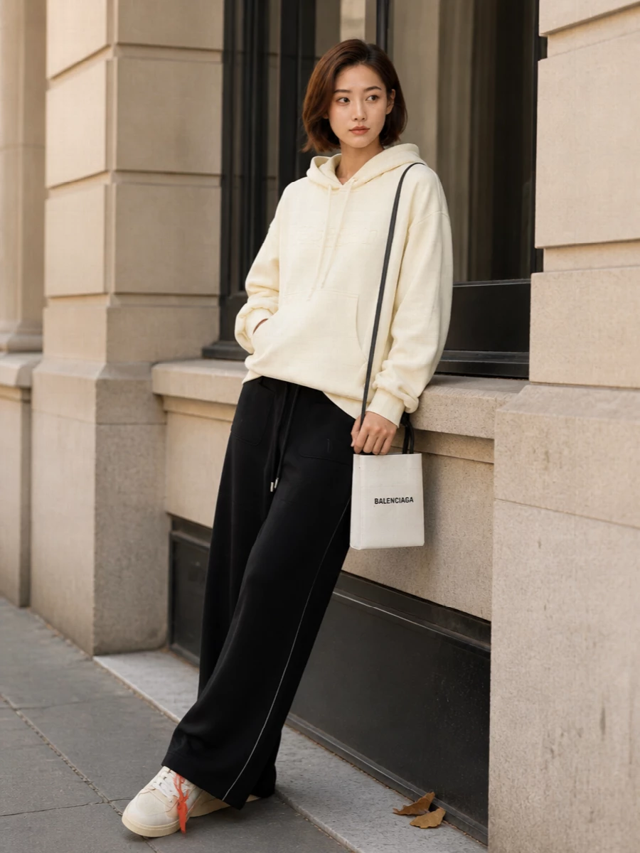 This look leans into elevated Off-White loungewear: the buttercream hooded knit and wide-leg black track pants form a relaxed, graphic base that lets the cream sneakers sit as a subtle focal point. The palette stays tight—creams, black, and a hit of red from the zip tag—while the Balenciaga mini tote sharpens everything with a structured, logo-forward finish. It’s an off-duty FW uniform that feels cocooning yet deliberately styled for the city.