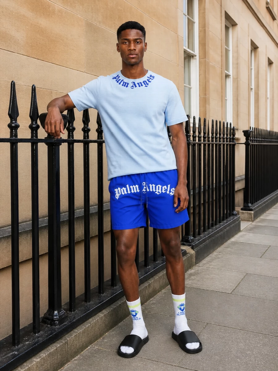 Electric blue shorts stay front and center against a clean Palm Angels palette, with the sky-blue tee creating a tight, tonal story up top. Black slides and backpack keep everything grounded, while the crisp white sport sock adds just enough color hit at the ankle to feel considered, not over-styled. It is an easy city-to-beach uniform that reads bold, graphic and relaxed all at once.