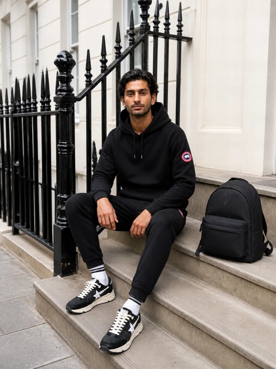 A monochrome Canada Goose set keeps the mood clean and athletic, with black tapered track pants mirroring the hoodie’s sharp, minimal energy. Chunky Golden Goose runners and a structured Moncler backpack push it firmly into city territory, more polished than pure gym kit but still easy to move in. The subtle striped sock is the only contrast, just enough to break up the black without diluting the streamlined feel.