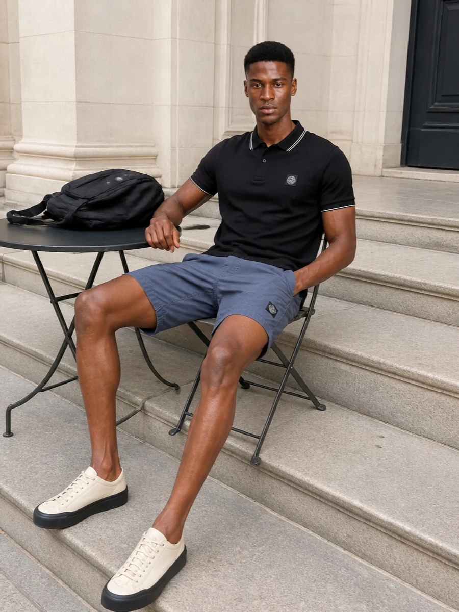 A black tipped polo and slate technical shorts create a clean, sport-driven summer uniform with a quiet hit of colour. Neutral, techy sneakers lift the palette without breaking the minimalist rhythm, while the backpack keeps the whole set firmly in the Stone Island street universe. It is sharp enough for the city, relaxed enough for coastal days and travel.