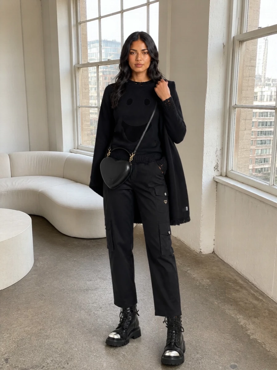 A head-to-toe black Moschino moment that leans into grungy streetwear: the cut-out smiley knit clashes playfully with sharp cargo pockets and heavy lug soles. The combat boots and mini heart bag keep the proportions short on top and strong at the base, so the attitude reads intentional rather than casual. It is a city-ready uniform for late nights, gallery runs, and anywhere you want your knitwear to carry the punch.
