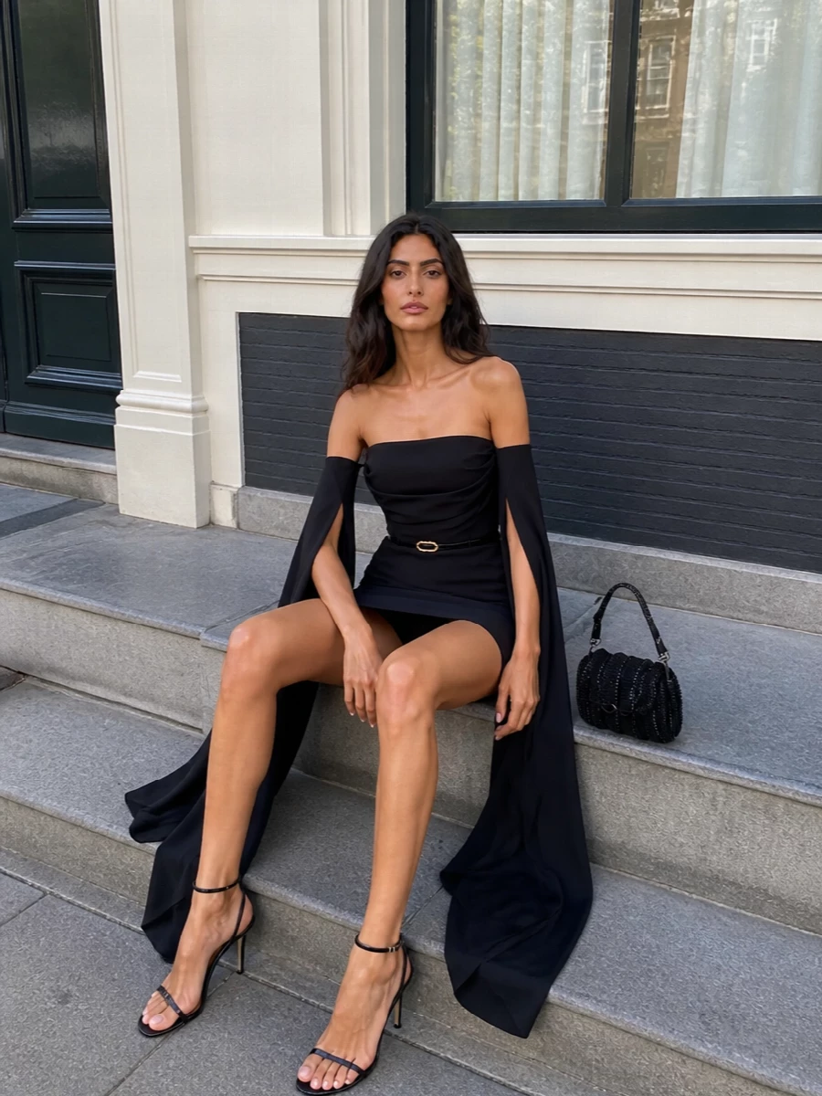 A pure-black, body-hugging mini is the canvas, while floor-grazing sleeves and a razor-sharp neckline bring full drama for night. Fine gold accents at the waist and on the heel stay controlled, and a jewel-like shoulder bag adds texture and movement without breaking the monochrome spell. The result is nightclub and late-dinner ready, with that photographed-from-every-angle intensity baked in.