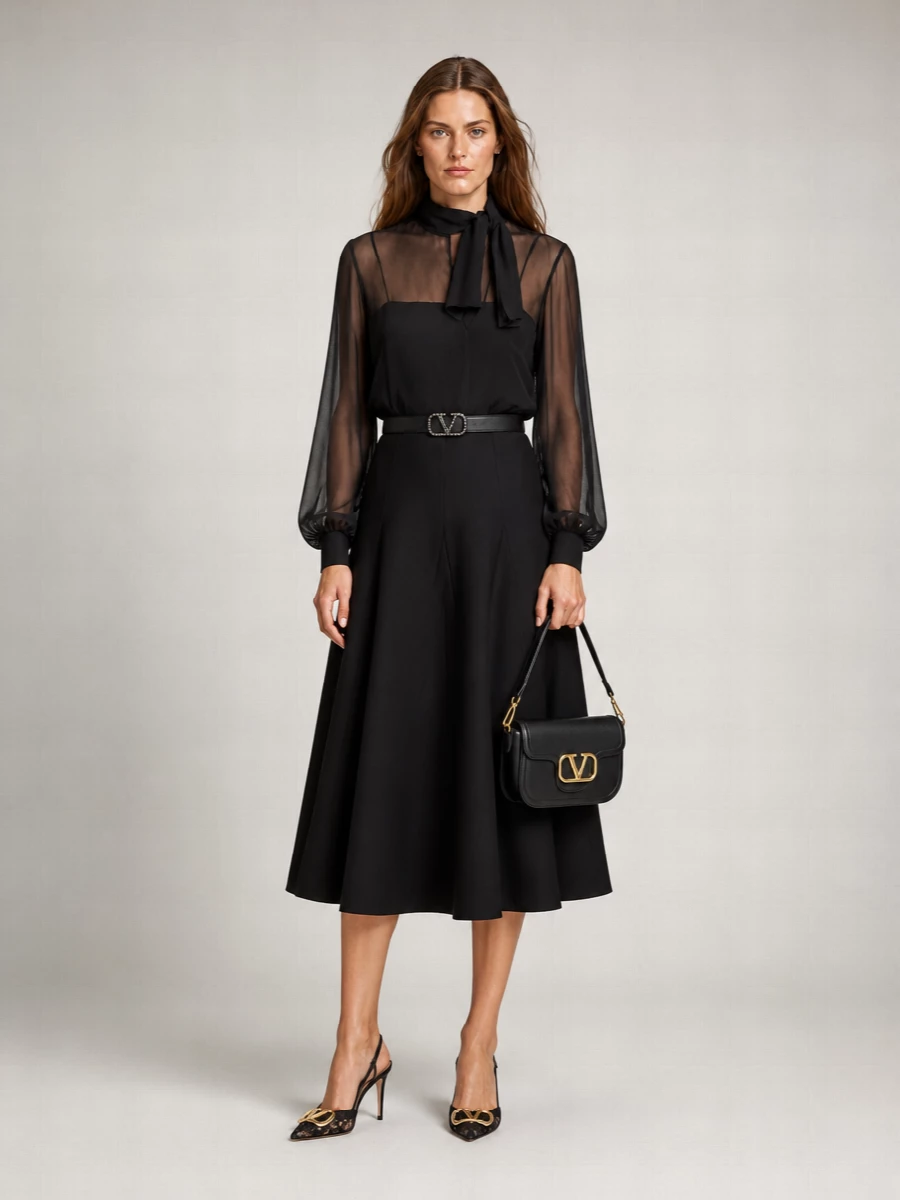 Sheer black romance sits over a sculpted flared midi, creating a long, fluid line that moves beautifully without feeling fussy. Lace-detailed slingbacks and a crystal-signed belt add just enough sparkle, anchored by a compact black shoulder bag that keeps the focus on cut and proportion. It is a quietly dramatic evening ensemble made for late dinners, gallery nights, and intimate events where detail reads up close.