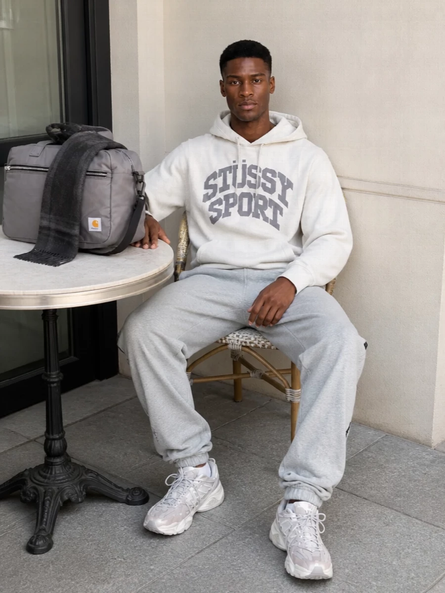 Off-white Stüssy Sport up top and heather grey track pants keep the outfit relaxed and clean, all in a cool neutral story. Chunky grey-and-cream runners echo the hoodie’s athletic feel while adding structure under the softer lines. A streamlined grey duffle and tonal tartan scarf round it out as a city-to-commute uniform that still feels sharp and considered.