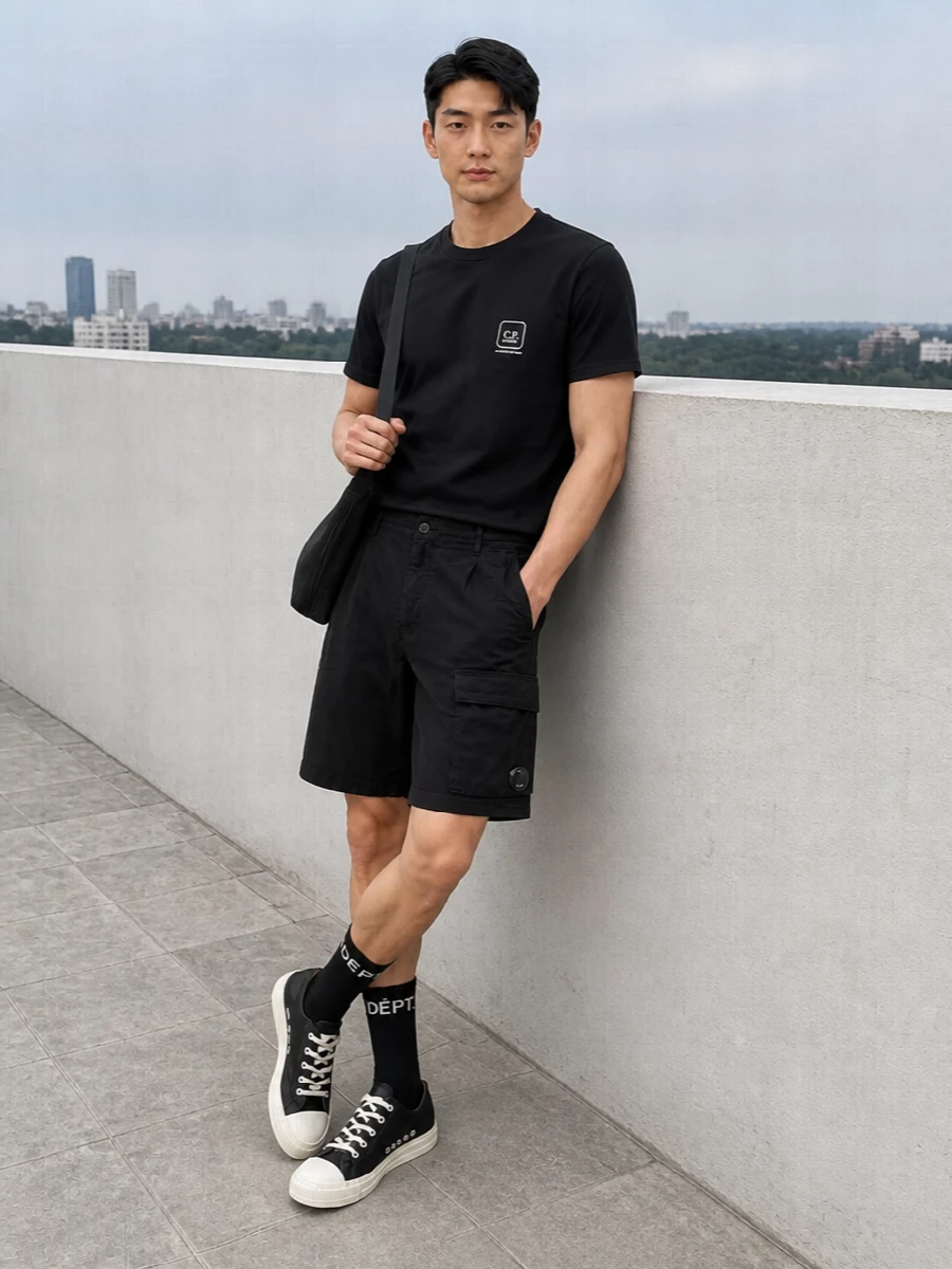 A strictly monochrome C.P. Company base keeps the focus on clean lines and subtle technical detailing, with black cargo shorts sharpening the way the tee reads. Rick Owens canvas sneakers and the pleated crossbody lean the outfit into quietly directional territory while staying very wearable. Logoed black socks finish it with just enough graphic attitude to feel intentional, not loud.