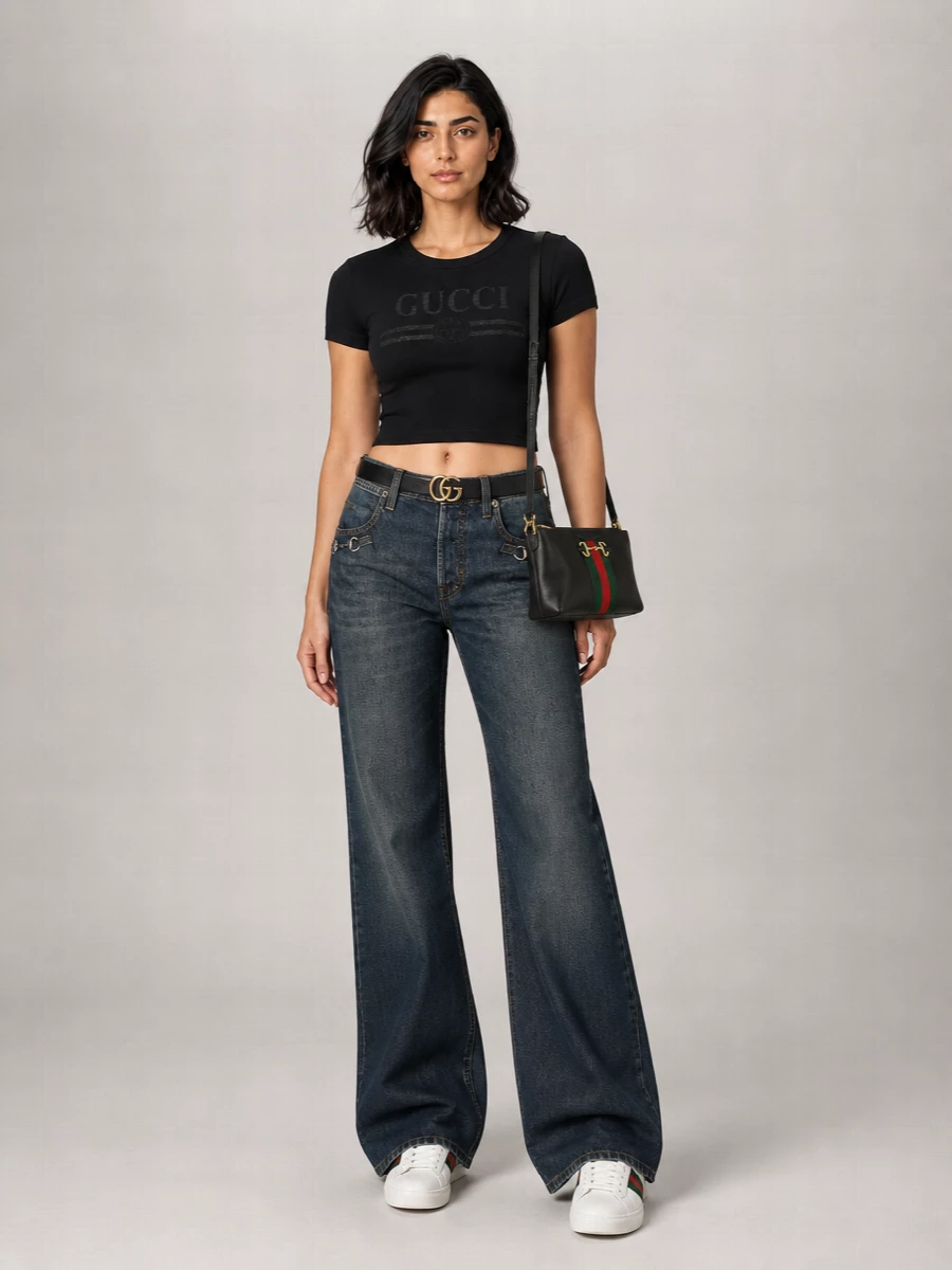 Gucci’s dark indigo wide-leg denim is paired with a cropped black logo tee for a clean, Y2K-inflected streetwear take that still reads polished. White web-detailed sneakers keep the line long and easy, while a slim black cross-body and matching belt echo the house hardware in a restrained way. The result is a streamlined, logo-aware Gucci ensemble that feels modern, graphic, and genuinely wearable all day.