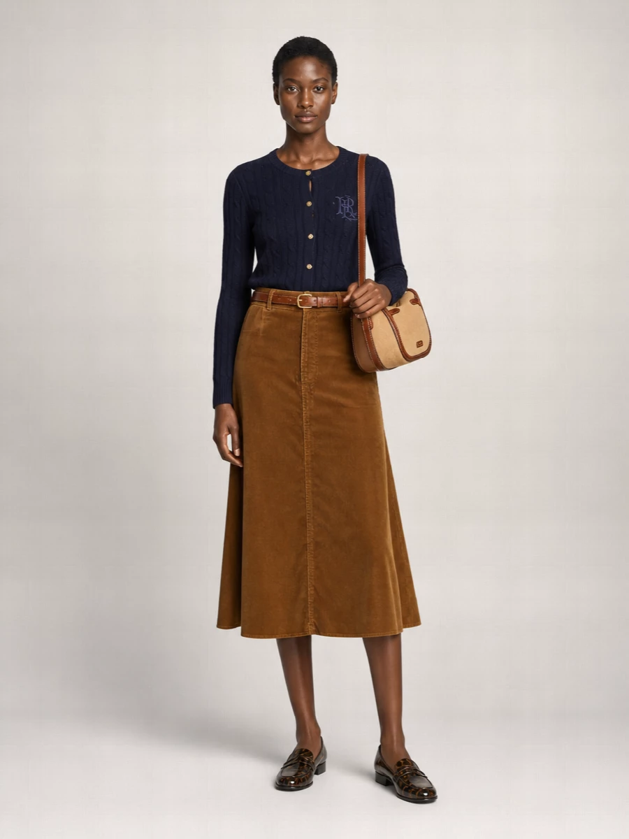 Navy and caramel play quietly against each other here, with the crest cardigan tucked into a swishy cord midi for a polished, collegiate line. Espresso loafers and a structured camel-and-chocolate shoulder bag ground the palette and keep it city ready. A slim tan belt is the final nod to equestrian ease, cinching the waist without shouting over the rest.