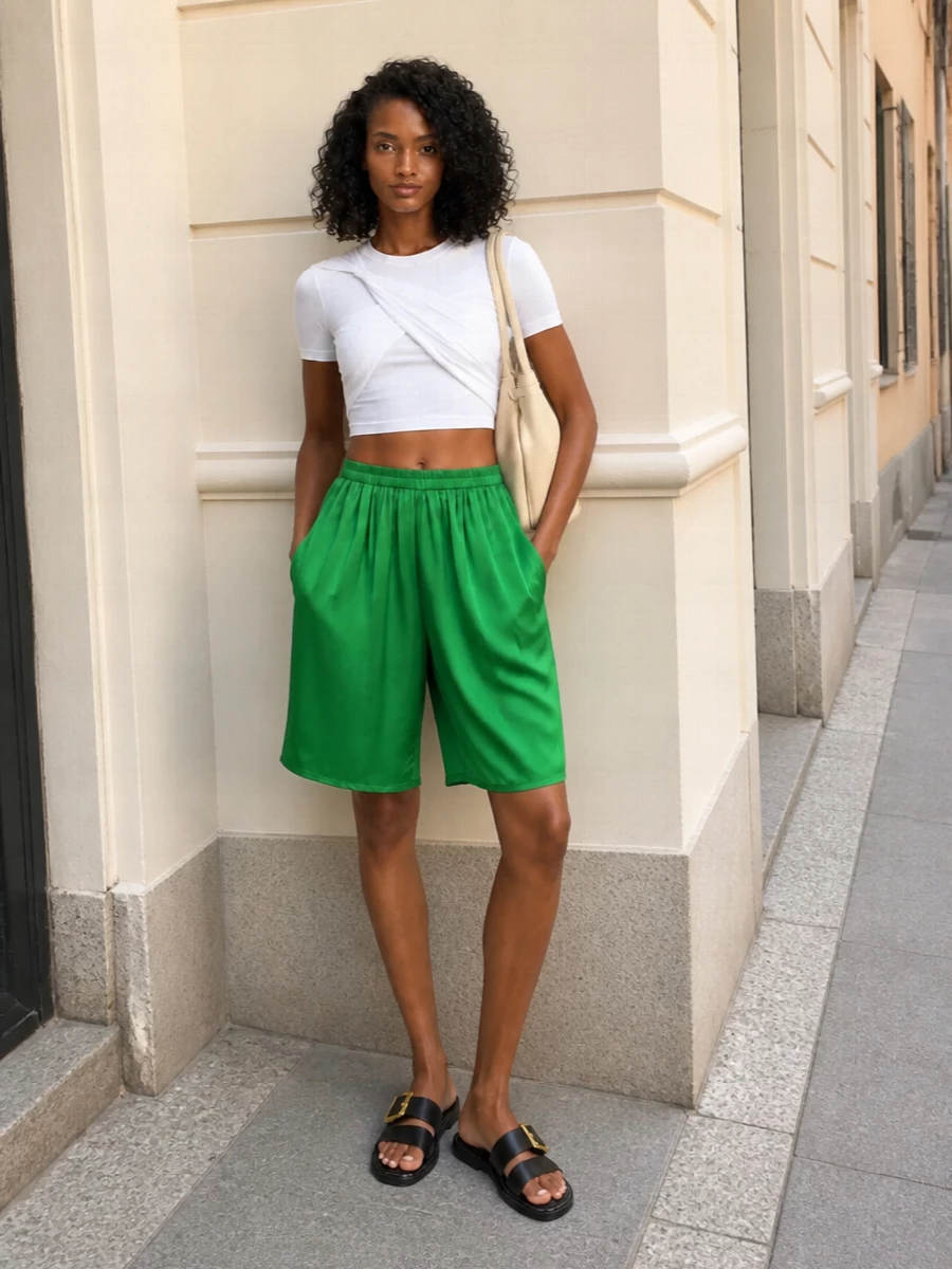 Liquid green satin meets a sculpted white crop for a sharp, minimal take on summer color. Black slides and a buttercream tote quietly ground the shine, keeping the palette clean and the mood relaxed but confident. The proportions stay modern by cinching the waist over easy, knee-grazing volume.
