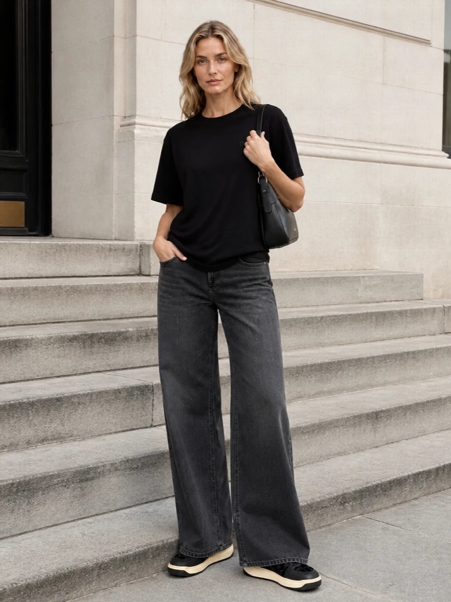 A longline black tee meets washed-charcoal wide-leg denim for a quietly graphic, street-focused silhouette. Black sneakers and a structured black shoulder bag keep everything in the same refined, low-key register, letting proportion and texture do the work. The result is relaxed but intentional, ideal for city days when you want polish without formality.