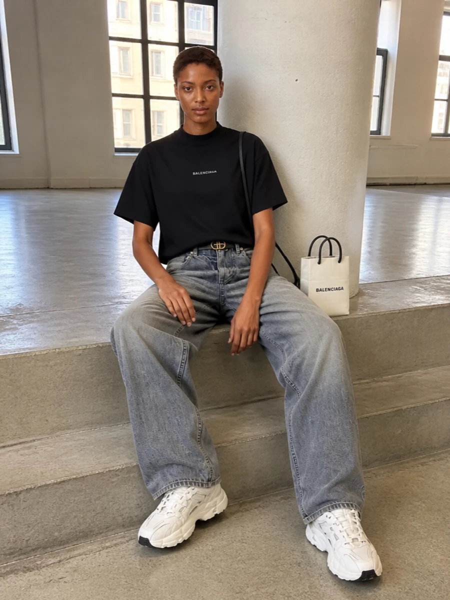 Balenciaga’s boxy black logo tee sits over washed grey wide-leg denim for a relaxed, long-line silhouette that feels intentional rather than sloppy. Chunky off-white sneakers and the sharp mini tote echo the black-and-white logo story, while the slim belt adds just enough structure and shine. It is an easy city uniform that reads sporty, graphic and quietly expensive all at once.