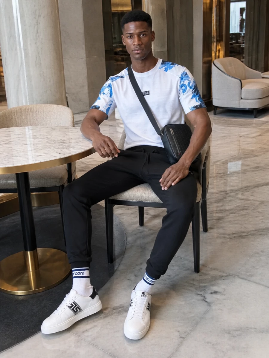 A graphic Versace tee meets streamlined black track pants for a clean, athletic base that keeps the blue marbling in the spotlight. White sneakers with a sharp black Greca band echo the logo story and keep everything feeling fast and modern. A compact black crossbody and striped sport socks finish it with just enough detail to feel thought through without losing that relaxed, off-duty ease.