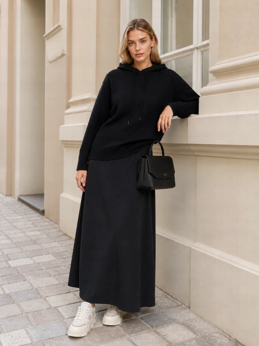 A black hooded knit meets a fluid charcoal maxi skirt for a quiet study in proportion, relaxed on top and long and clean through the line. Soft off‑white sneakers keep it grounded in the street while a structured black bag adds just enough city precision. The result is easy, monochrome and quietly confident for days that move between errands, desks and late coffees.